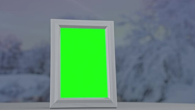 Empty photo frame with green chromakey stands on windowsill with view of winter nature through window glass outdoor