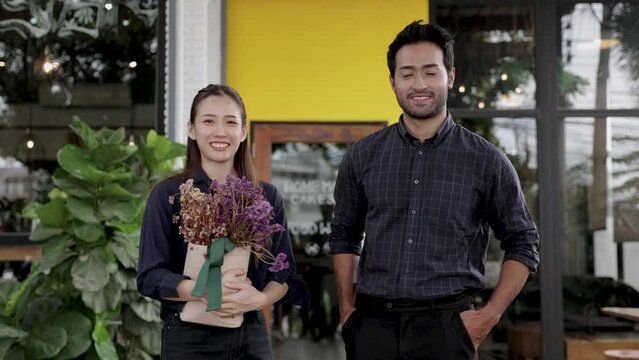 Two cheerful baristas multiethnicity man and woman running coffee shop business entrepreneurship standing in front of cafeteria with flowers smiling, Asian and Indian couple small business strat up