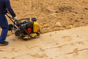 Worker using vibratory plate compactor for compaction sand during path construction.