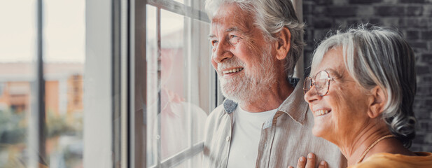 Happy bonding loving middle aged senior retired couple standing near window, looking in distance,...