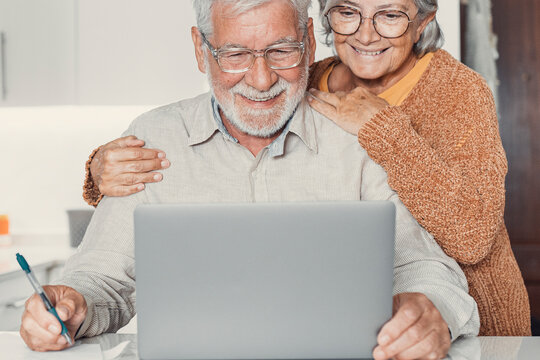 Old age and modern tech. Retired family couple sit at kitchen table use laptop watch photo video read latest news online. Aged husband share discuss interesting information on website with senior wife