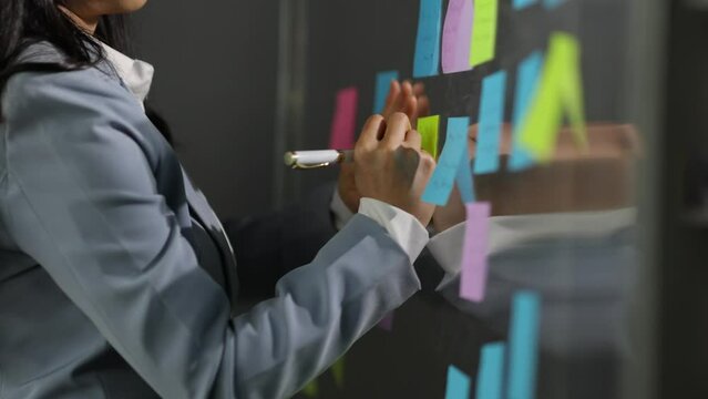 Creative young Asian woman thinking and planning a startup business with paper stickers on a transparent board.