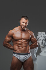 Fototapeta premium Portrait of a smiling, rugged man with a well-groomed model appearance, showcasing his bare, perfect muscular torso while standing next to ancient Greek statues on a gray