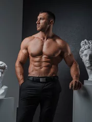 Zelfklevend Fotobehang Portrait of a rugged man with a well-groomed model appearance, dressed in classic trousers with a bare muscular torso, posing next to ancient Greek statues on a gray background © Fxquadro