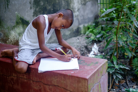 South asian primary school going student reading and writing in a white notebook sitting in front of his home 