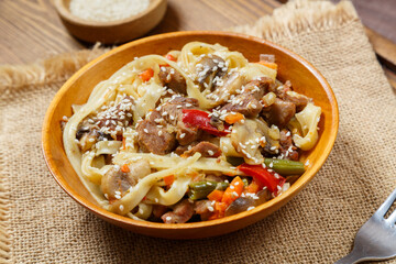 Wok udon noodles with beef and vegetables in a wooden plate on a napkin with a fork next to sesame seeds.