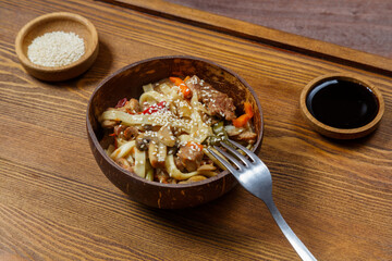 Wok udon noodles with beef and vegetables in a coconut shell plate on a napkin with a fork next to...