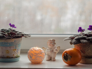 Horizontal close up of a white windowsill with two pots of blooming violets, a candle lit, a...