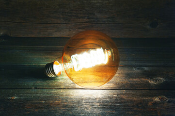 A decorative burning light bulb lies without a connection on old wooden boards in the dark. Side...