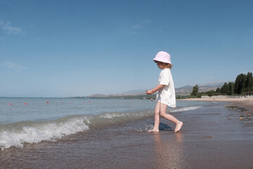 A little fair-haired girl in a Panama hat and white clothes is running along the seashore. Concept of safe water games, healthy lifestyle.