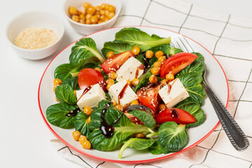 Fresh pak choi salad with tomatoes, chickpeas and feta cheese toping with sauce and sesame seeds in a plate with a fork on checkered napkin