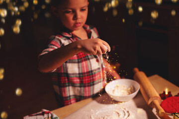 A little boy in red checkered pajamas sips flour on the table while preparing Christmas gingerbread...