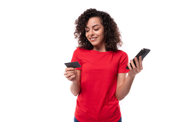 Naklejka premium young pretty curly brunette woman dressed in a red basic t-shirt enjoys online payments. e-business concept