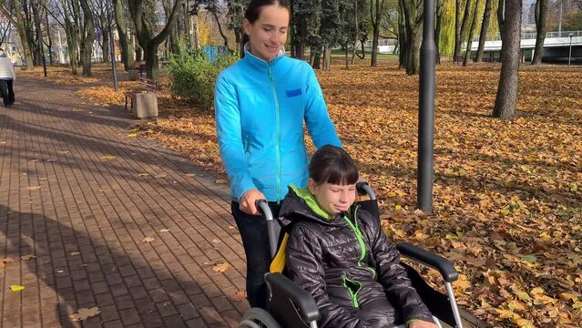 Mom with her little disabled daughter in a wheelchair in park. Happy mother and child hugging and love in family