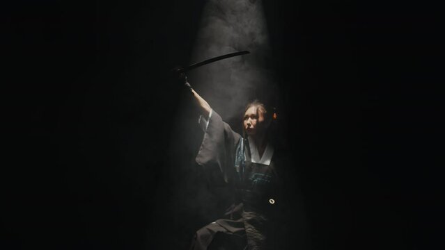 Young asian woman in a kimono standing in a dark studio under the beam of light, takes out a katana from its sheath and raises it up among a streaming smoke