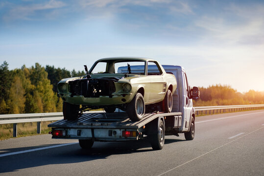 Fototapeta A Towing Truck In Motion With A Damaged Car After The Traffic Accident On A Road. Emergency Roadside Assistance. Retro Car Project Renovation. Vintage Car Recovery And Reanimation.