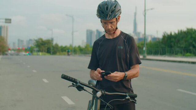 Tilt up shot of man in sportswear and helmet standing by bicycle on road, using map on smartphone and looking around