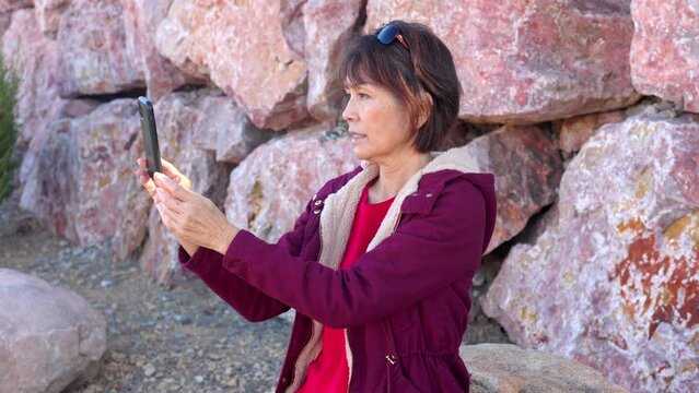 Attractive middle age woman sitting on bench on outdoors hike taking photos with smartphone smiles looking at pictures. Mature confident mature asian lady enjoying walk sitting near red rock wall.