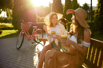 Two gorgeous women in sunlight sitting on bench chatting drinking coffee