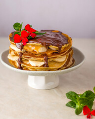 Pancake cake drizzled with chocolate on a stand decorated with mint and a red flower, close up.