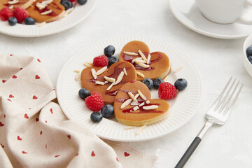 A heap of heart shaped pancakes with berries and a fork on the white plate on grey background
