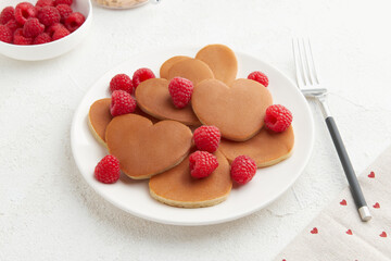 A heap of heart shaped pancakes with raspberries and a fork on the white plate on white background