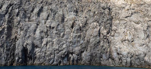 Weathered seaside rock face texture with parts of green and blue water. Aged volcanic stone wall surface background pattern with cracks and scratches. Banner. Ischia Island, Italy.