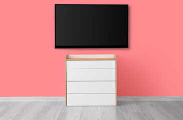 Modern smart TV set hanging on pink wall near chest of drawers in room