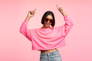 Asian woman holding hands up pointing fingers wearing sunglasses dancing isolated on pink background