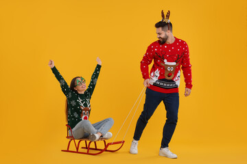 Young couple in Christmas sweaters. Man pulling his woman in sled on orange background