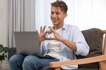 Happy young man having video chat via laptop and making heart with hands on armchair indoors. Long-distance relationship