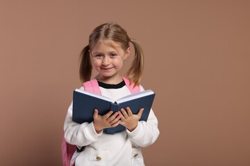 Happy schoolgirl with backpack and book on brown background