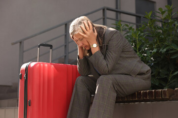 Being late. Frustrated senior businesswoman with red suitcase sitting on bench outdoors