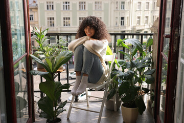 Beautiful young woman relaxing in chair surrounded by green houseplants on balcony - Powered by Adobe