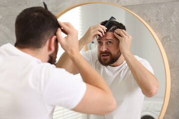 Dandruff problem. Man with comb examining his hair and scalp near mirror indoors