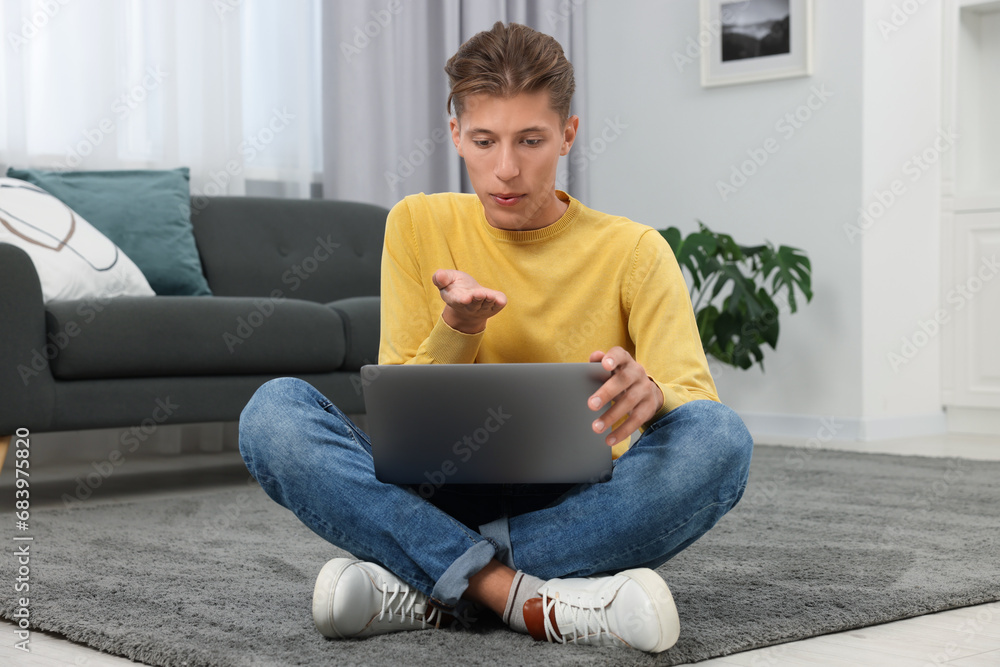 Poster Happy young man having video chat via laptop and blowing kiss on carpet indoors. Long-distance relationship