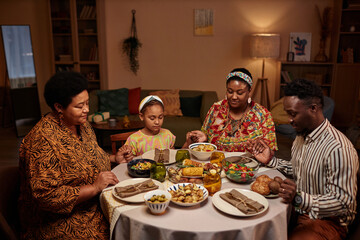 Black family sitting at Kwanzaa dinner table with eyes closed, hold hands and praying