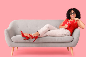 Beautiful woman in 3D glasses with soda on sofa pointing at viewer against pink background