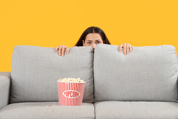 Young woman watching movie behind sofa on yellow background