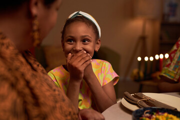 Cute excited preteen girl looking at mother and covering mouth when laughing at family dinner