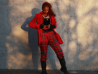 Selfie photo of a woman near a white gray wall in tight black clothes, a red raincoat, a leather bag. Cinematic punk style, fashion.