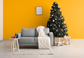 Beautiful Christmas tree with gift boxes and sofa near orange wall