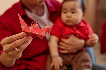 Close up of caring Asian father with cute little baby holding red paper crane to camera as symbol...