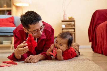 Front view of happy Asian father playing with cute baby boy lying on floor at home and holding red...