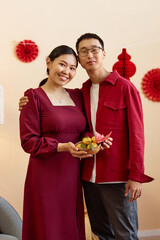 Vertical portrait of young Asian couple looking at camera and wearing red indoors while celebrating...