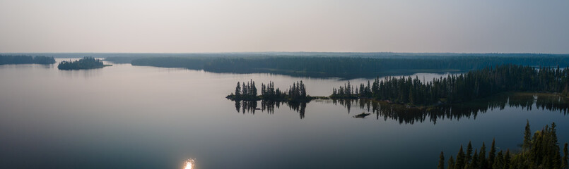 Aerial view of a large calm northern lake with islands and points of land. The forest is heavily treed with pine and spruce. The sun and sky are reflecting on the surface of the water.  The atmosphere