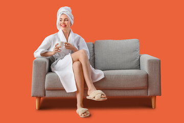 Young woman with coffee cup after shower sitting on grey sofa against orange background