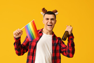 Young man in reindeer horns with LGBT flag and champagne on yellow background