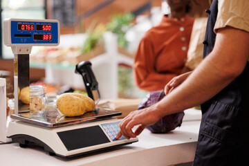 Diligent seller in a small grocery store measures the weight of organic potatoes using an electronic scale. Caucasian man employed as cashier assisting a customer in weighing locally grown potatoes.