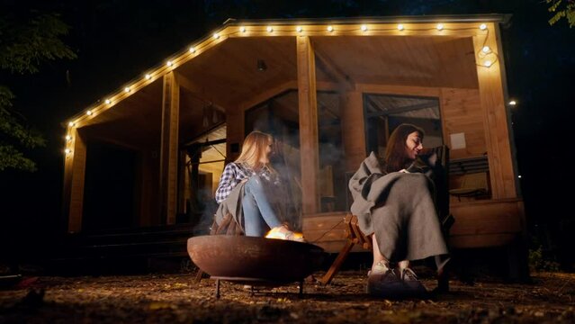 shot of girl friends sitting together outside at night by fire warming their hands and chatting outside their wooden house